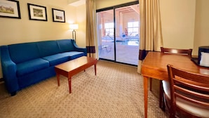 Habitación de hotel con un sofá azul, una mesa de madera y una mesa de comedor con sillas. Se ve un ventanal grande con cortinas, y un cuadro en la pared.