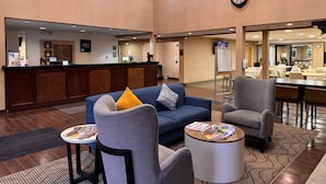 Un vestíbulo de hotel con mostrador de recepción, zona de estar con sofás y sillones, un reloj en la pared y una pantalla de televisión.