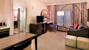 A hotel room with a desk, a chair, a sofa, a TV, a mirror, and a window with curtains.