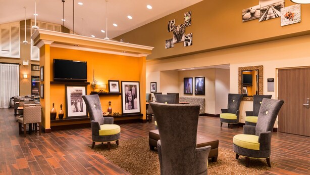A hotel lobby with a wooden floor, grey upholstered chairs, a flat-screen TV, framed pictures on the wall, and a reception desk.