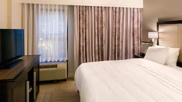 A hotel room with a large bed, a flat-screen TV on a wooden dresser, a window with sheer curtains, and a patterned curtain.