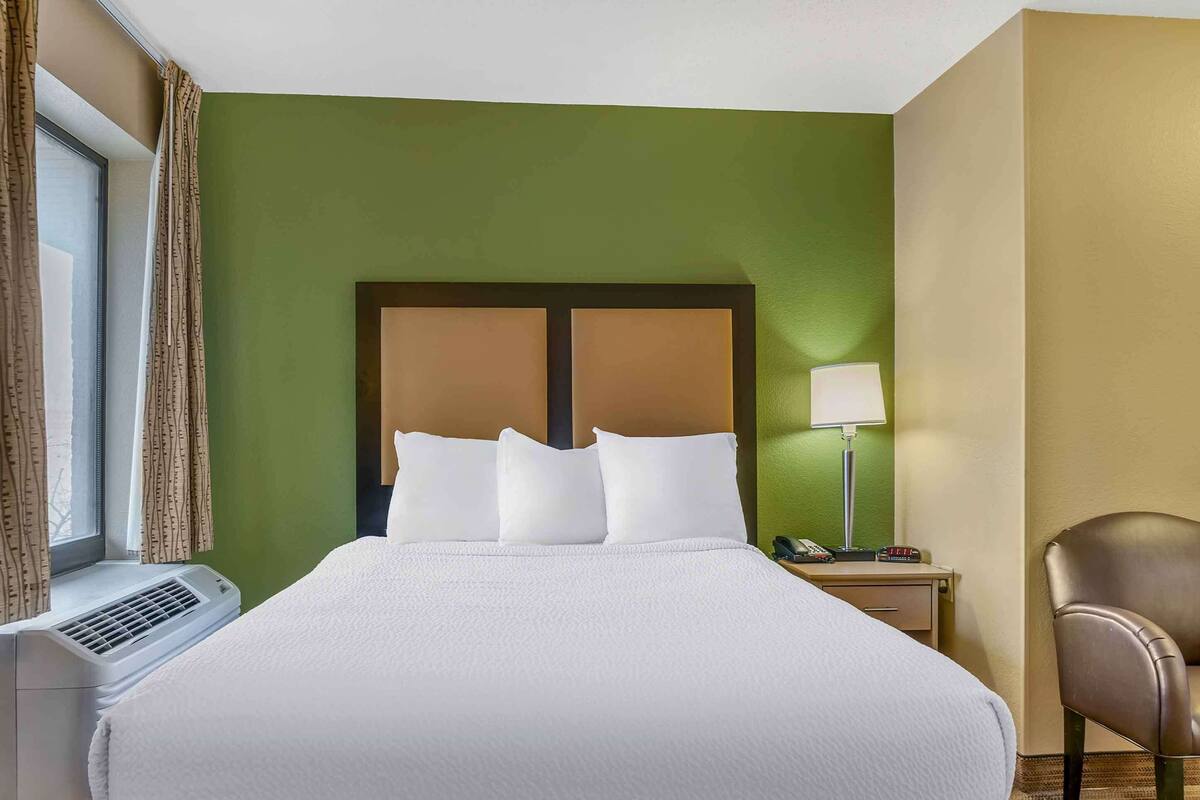 Habitación de hotel con una cama grande, una lámpara de noche, una silla y una ventana con cortinas.