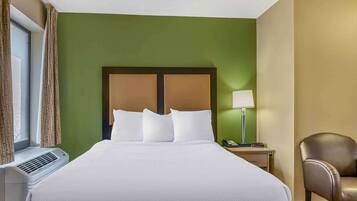 Quarto de hotel com uma cama grande, luminária de cabeceira, uma cadeira e uma janela com cortinas.