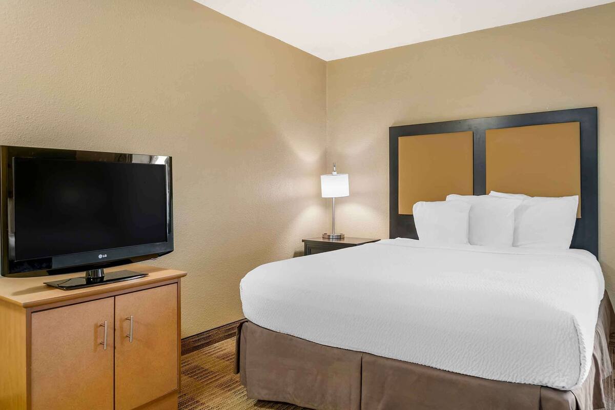 Habitación de hotel con televisión de pantalla plana sobre un mueble de madera, una lámpara de noche y una cama con cabecero.