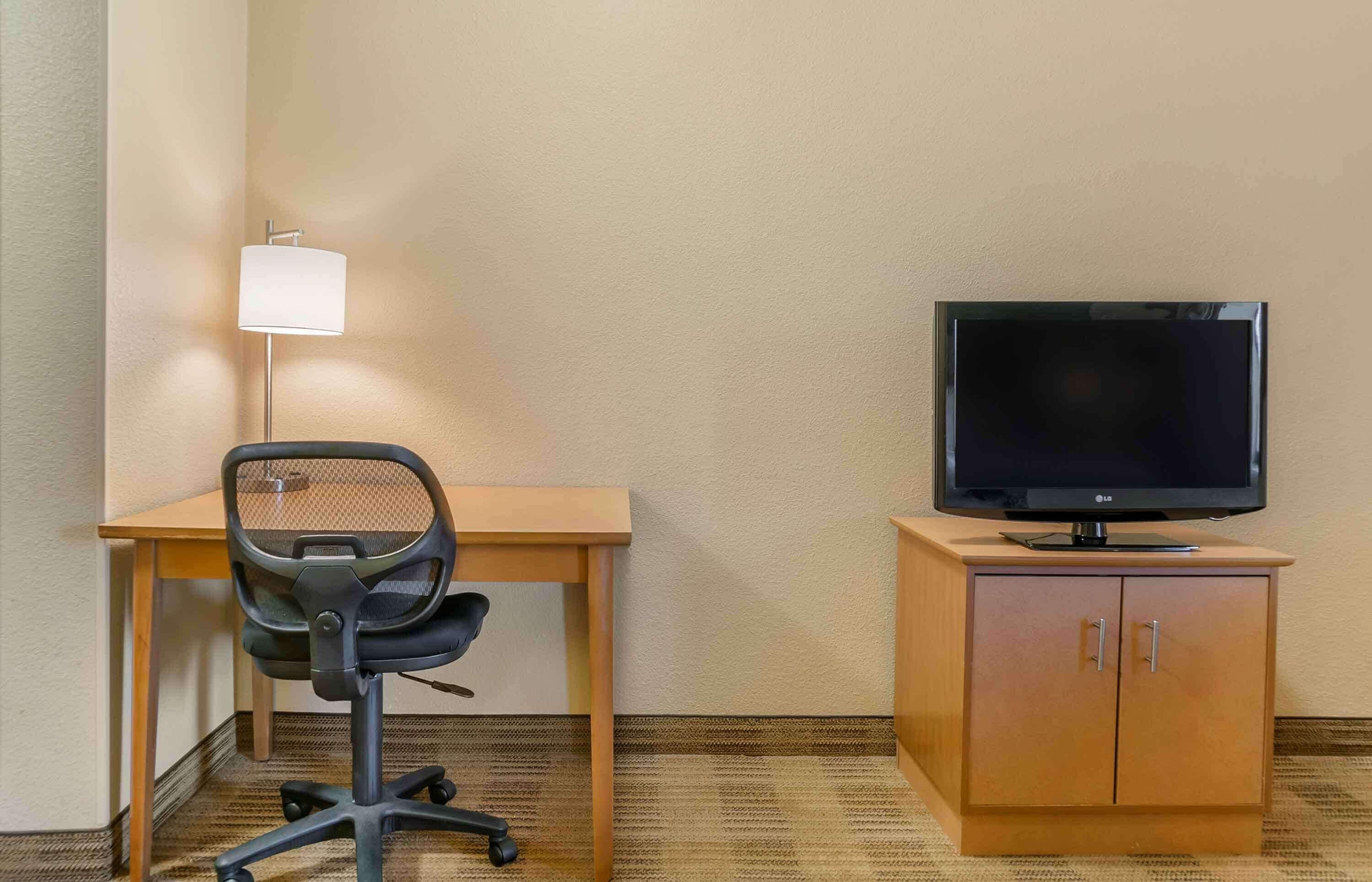 Une chambre d’hôtel avec un bureau en bois, une chaise de bureau noire, un téléviseur posé sur une commode en bois et une lampe sur pied.