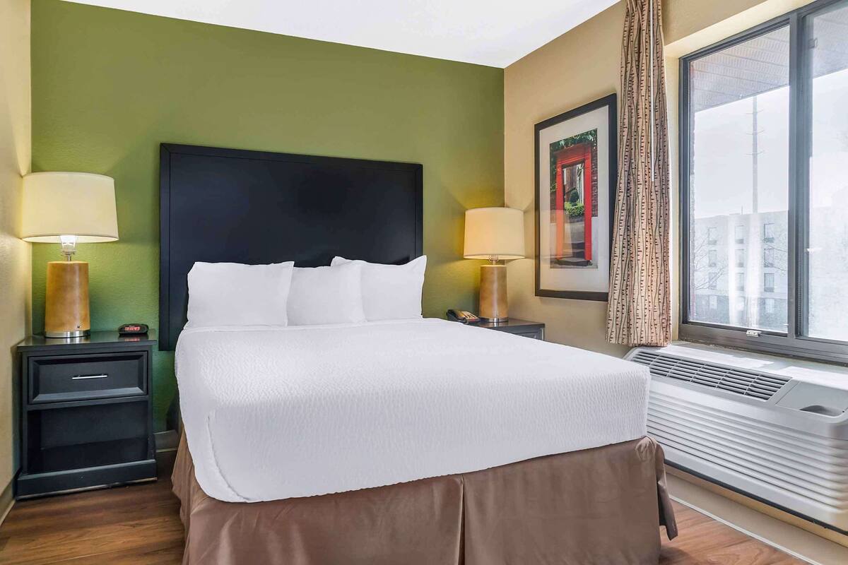 Una cama bien hecha en una habitación de hotel con paredes verdes, dos mesitas de noche con lámparas, un cuadro enmarcado en la pared y una ventana con cortinas.