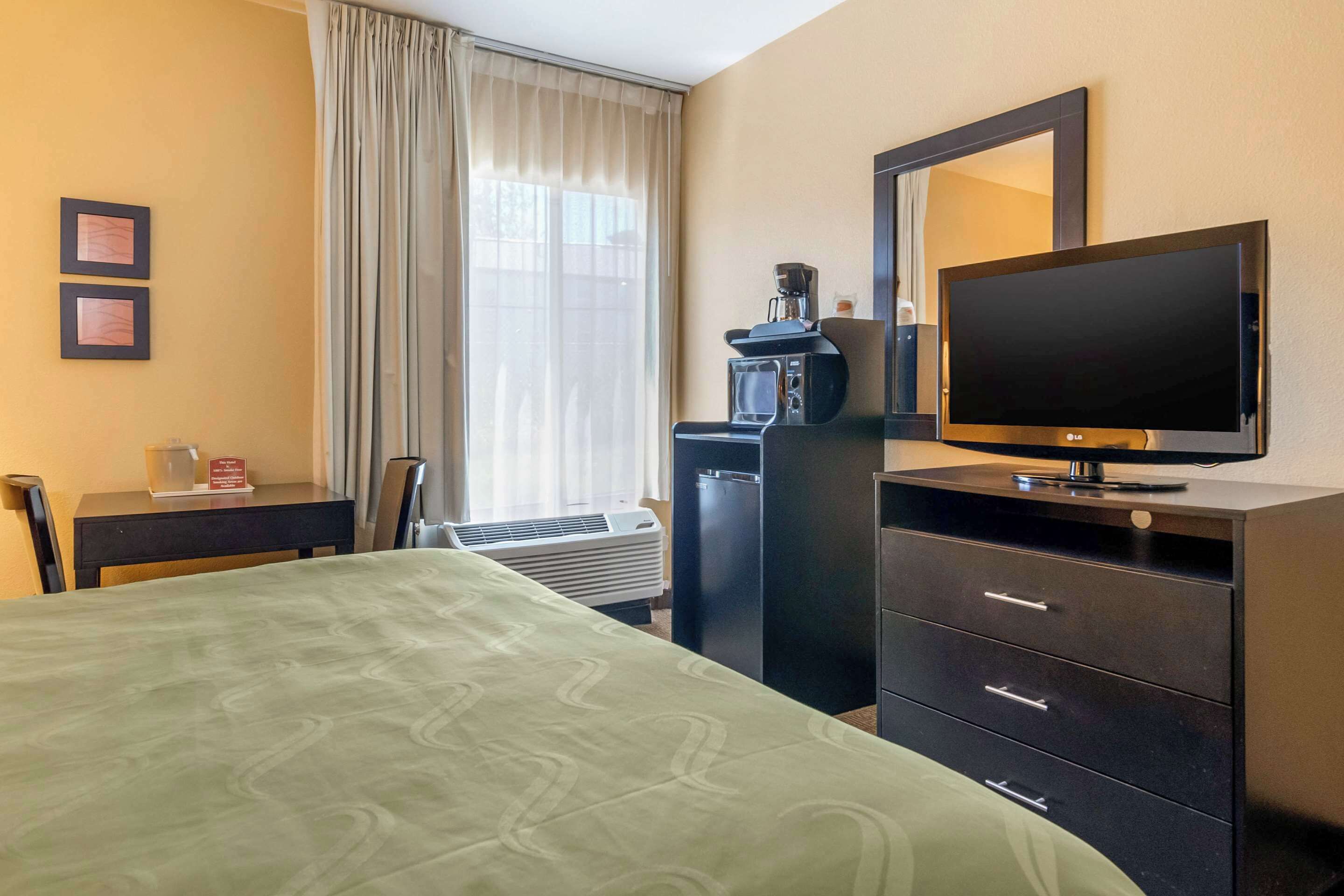 Quarto de hotel com uma cama, escrivaninha, televisão, cômoda e janela com cortinas.