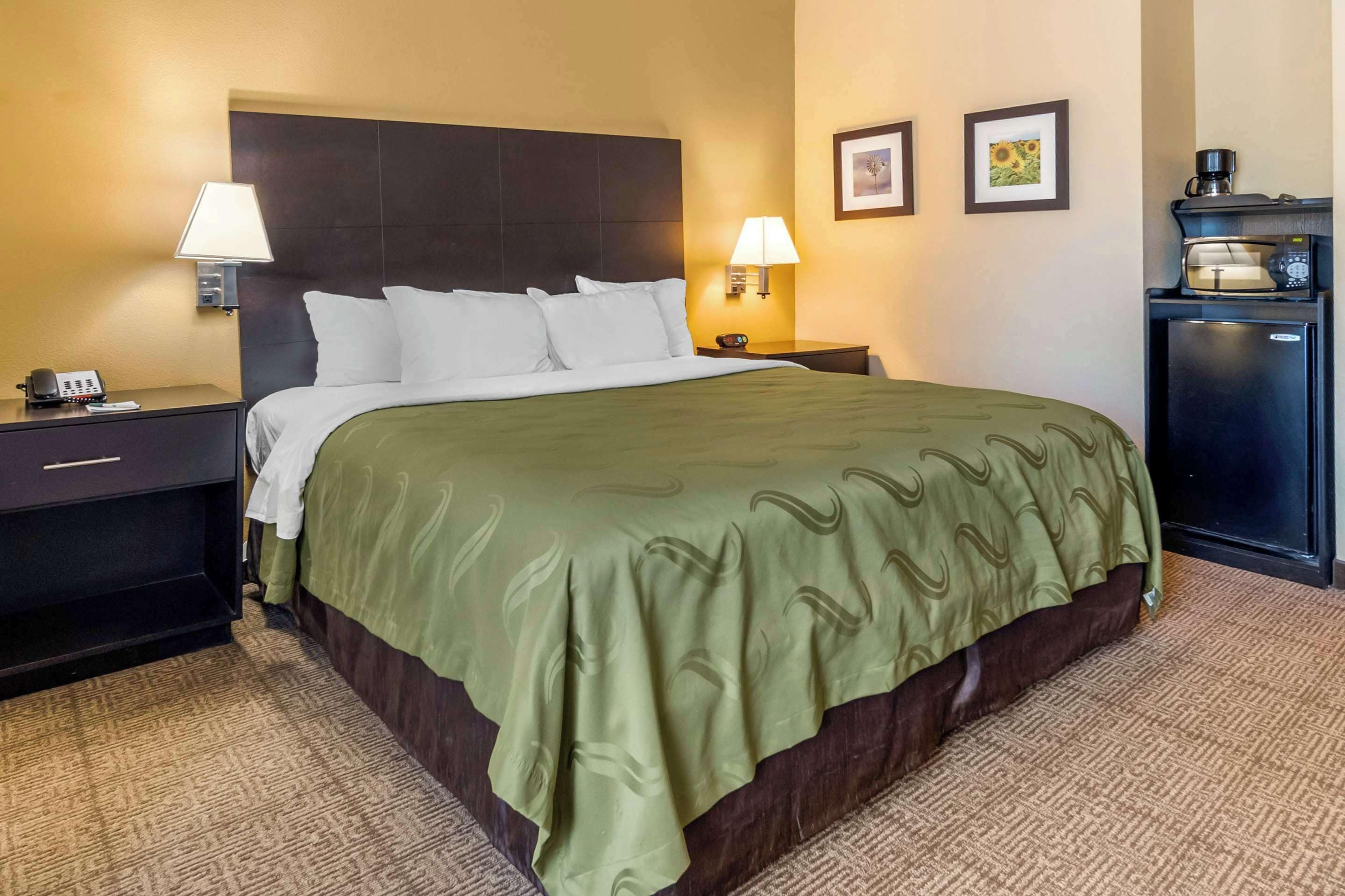 Habitación de hotel con una cama grande, dos lámparas de noche, una mesita de noche con teléfono, una pequeña mesa con cafetera y un refrigerador.