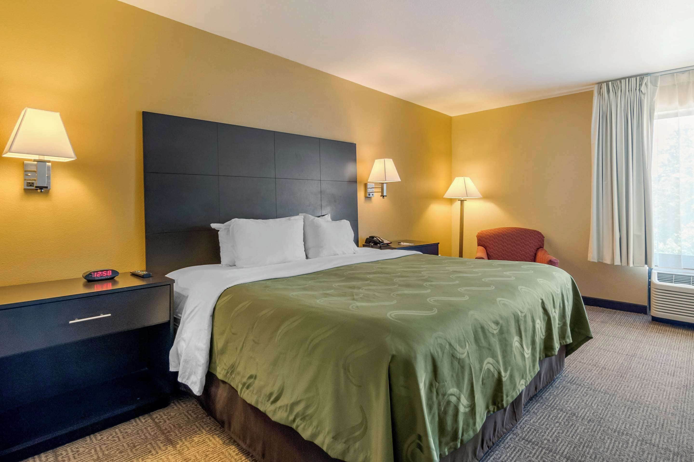 Habitación de hotel con una cama grande, dos lámparas de noche, una mesita de noche con un reloj y una ventana con cortinas.