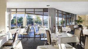 A restaurant with tables set for dining, large windows offering a view of the outdoors, and a potted plant on the right side. - Garden City Hotel, BW Signature Collection (Narrabundah)