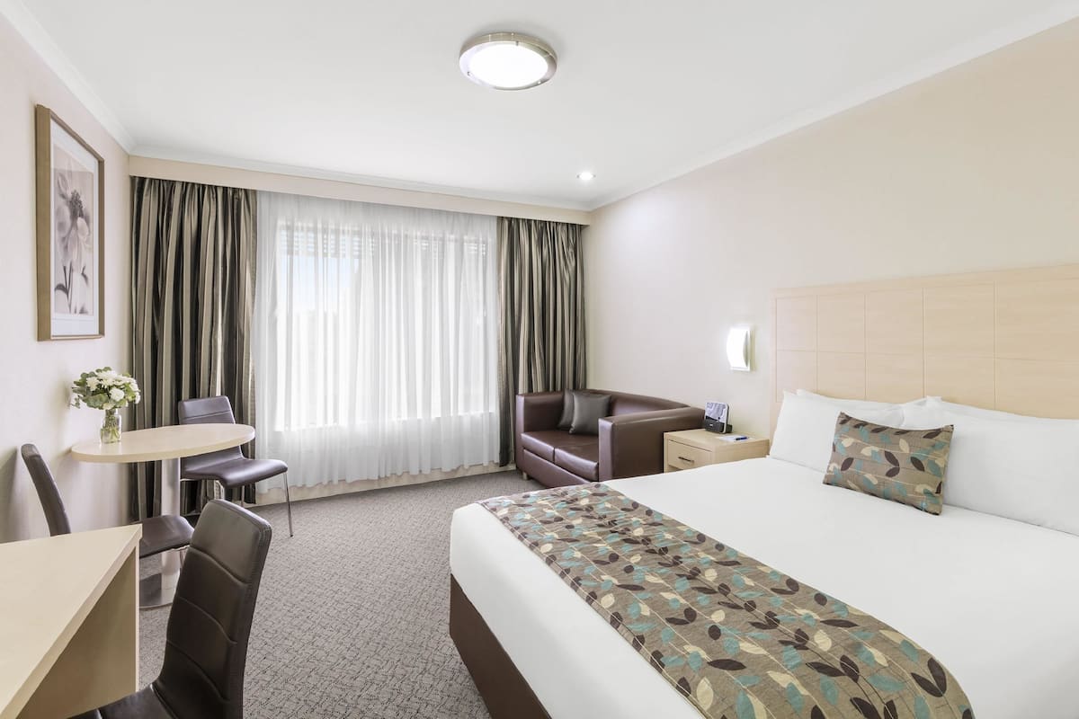 A hotel room with a bed, a desk, a chair, a small table with a vase, and a window with curtains.