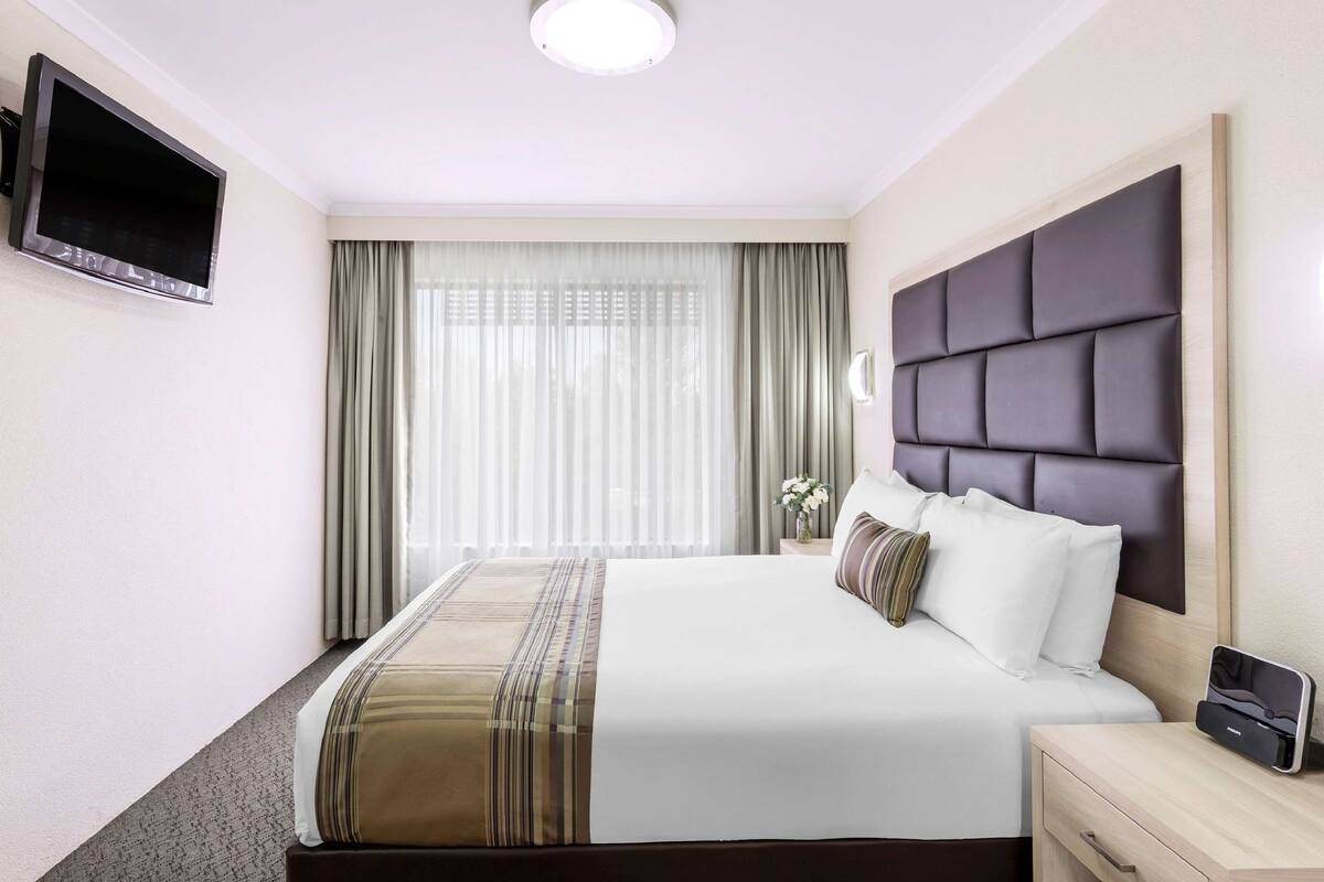 A hotel room with a large bed, a flat-screen TV mounted on the wall, a bedside table with a lamp, and a window with sheer curtains.