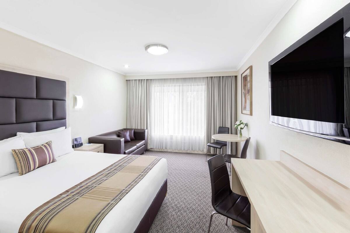 A hotel room with a large bed, a desk, a chair, a sofa, and a TV mounted on the wall.