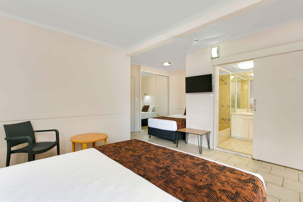 A hotel room with a bed, a desk, a chair, a small table, a TV, and a bathroom visible through an open door.