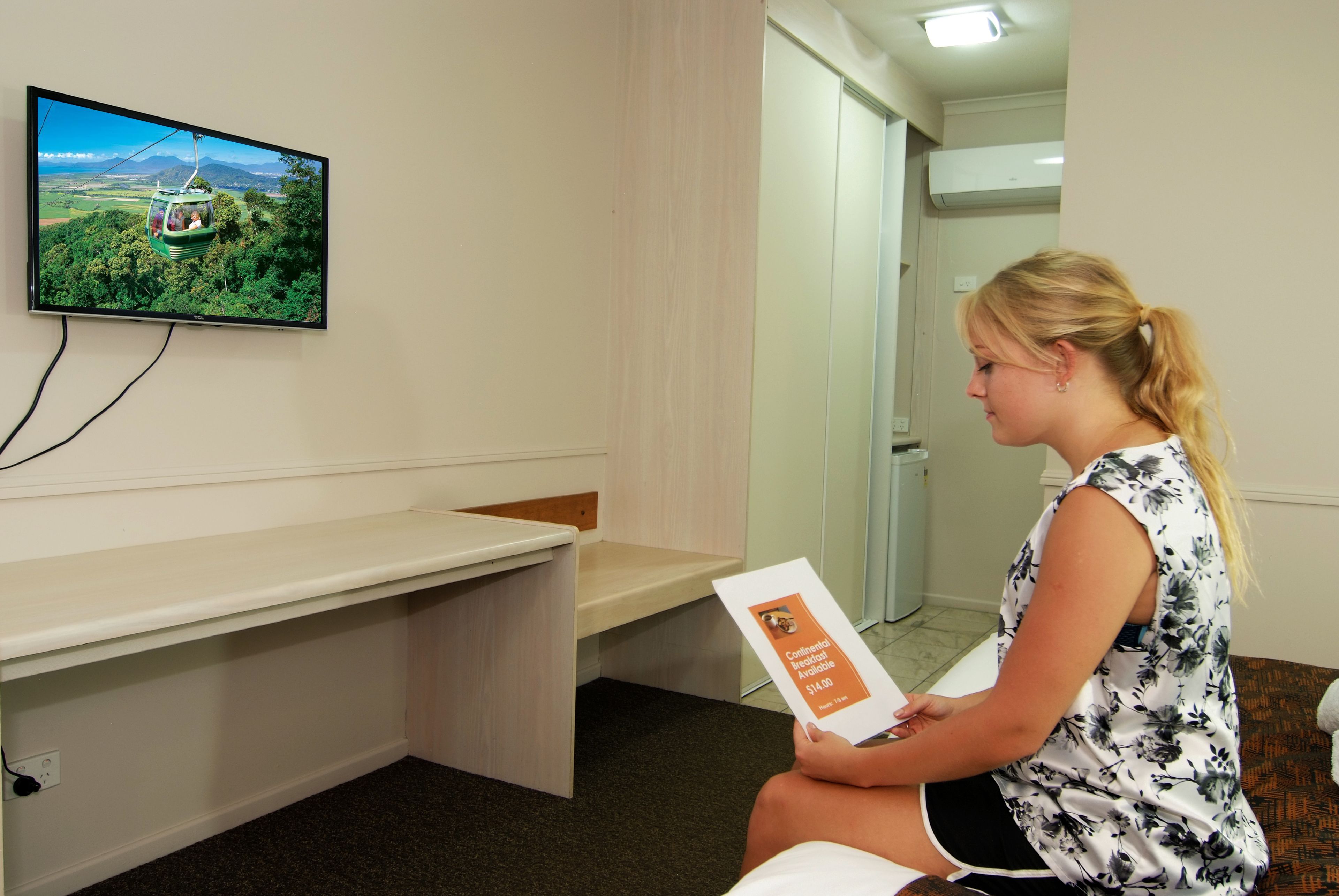 A woman sitting on a bench in a room with a television displaying a scenic view, a sign with a price, and a door in the background.