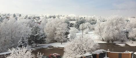 A snow-covered outdoor area with a parking lot, a red vehicle, and a building with a balcony.