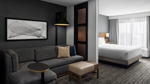A modern hotel room with a grey sofa, a round side table, a wooden bench, a bed with white linens, a black floor lamp, a framed abstract artwork, and a large window with sheer curtains.