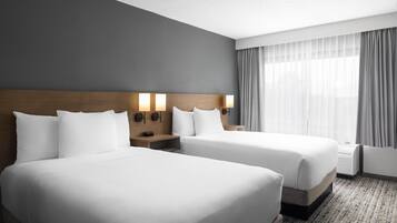 A hotel room with two beds, a wooden headboard, bedside lamps, a large window with curtains, and a carpeted floor.