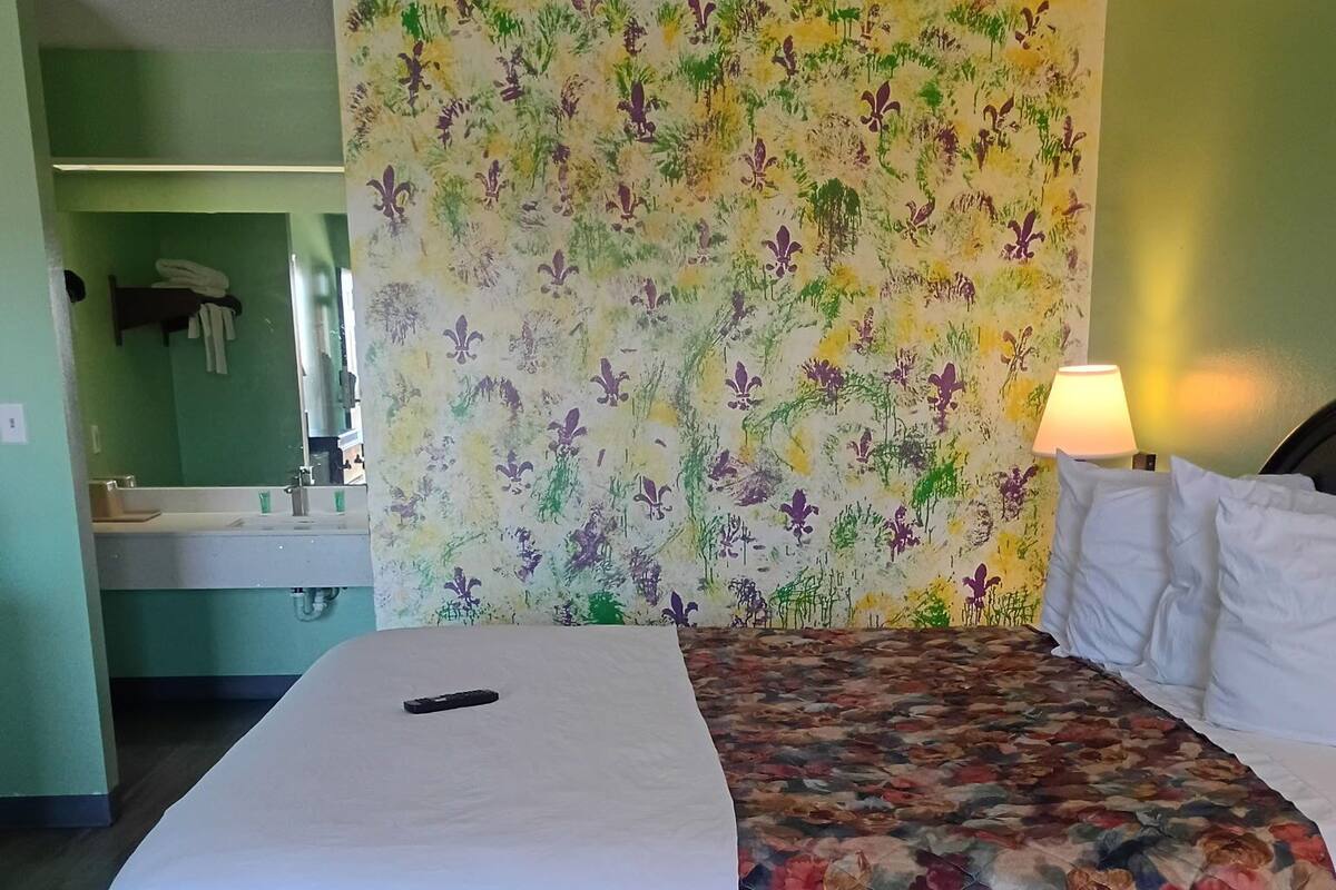 A hotel room with a bed, a floral-patterned headboard, a bedside lamp, and a bathroom visible in the background.