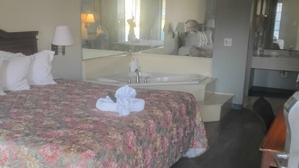 Una habitació d'hotel amb un llit, una taula de treball, una cadira, un bany amb un lavabo i un mirall, i un mirall gran a la paret.