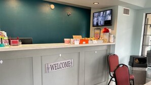 Una zona de recepció amb un cartell que diu 'WELCOME', un taulell amb diferents objectes, una pantalla de televisió i dues cadires vermelles.