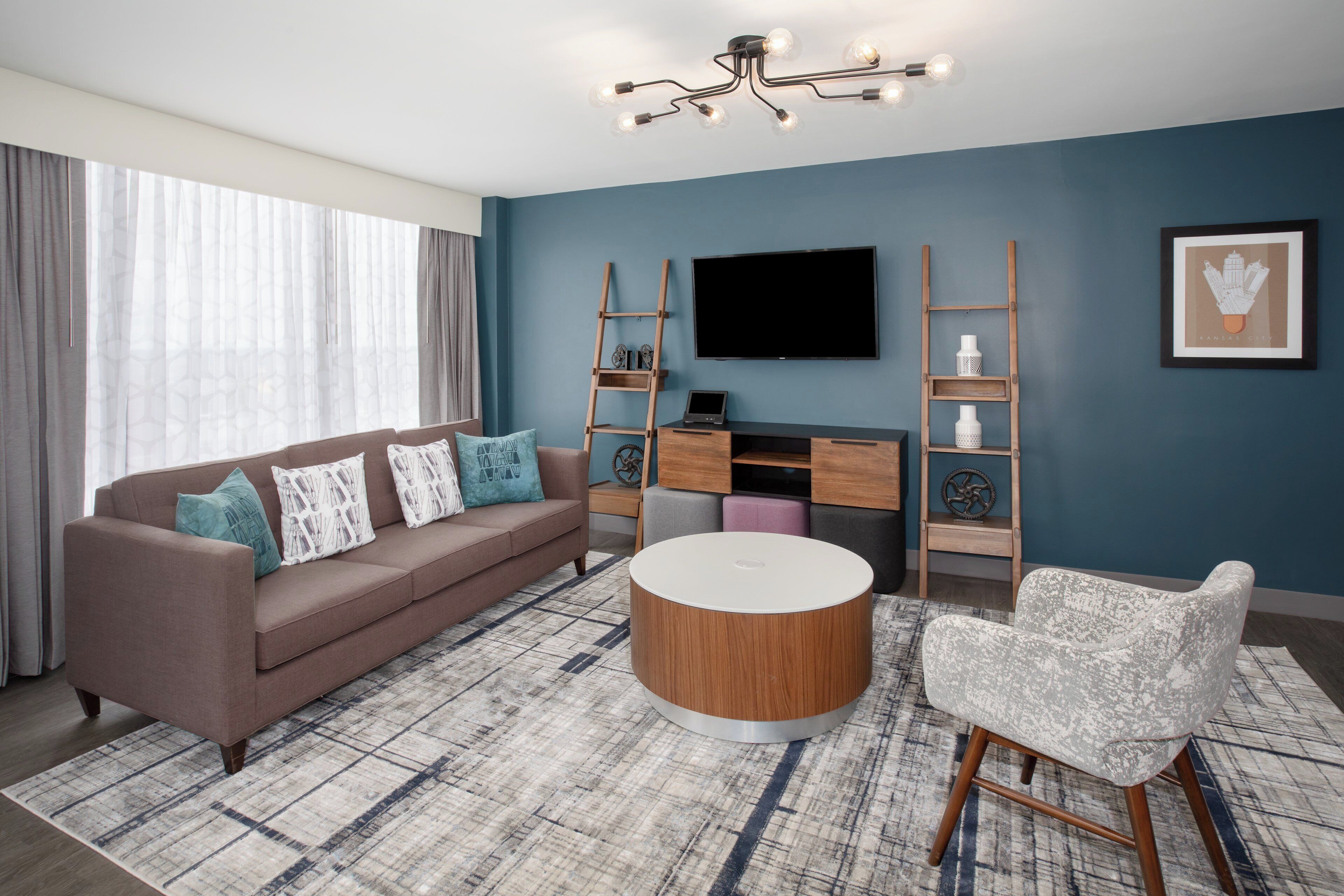 Una sala de estar moderna con un sofá, una mesa de centro y dos sillones. Un televisor de pantalla plana está montado en la pared, y hay estanterías de madera con objetos decorativos.