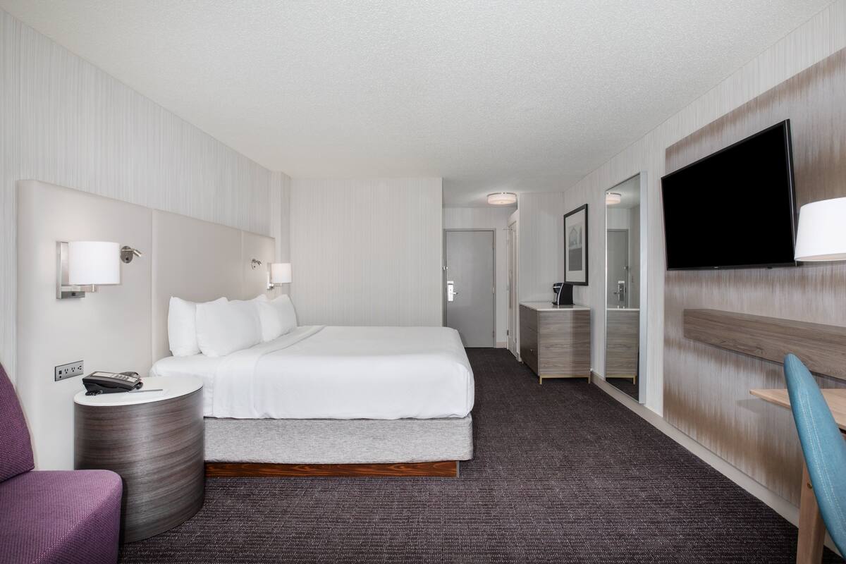 Habitación de hotel con cama, mesita de noche con lámpara, televisor montado en la pared y un pequeño rincón de descanso con una silla.