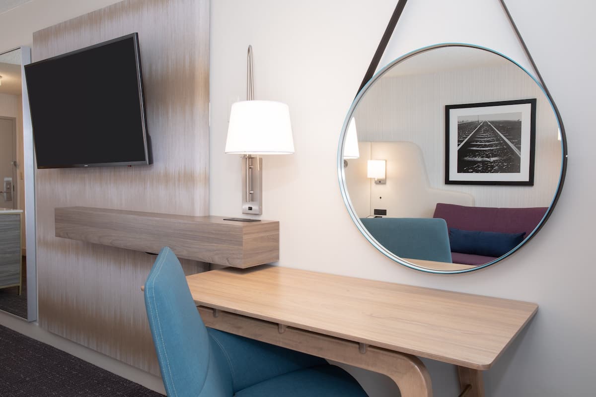 Habitación de hotel con televisión de pantalla plana, escritorio con silla, espejo redondo y un cuadro en la pared.