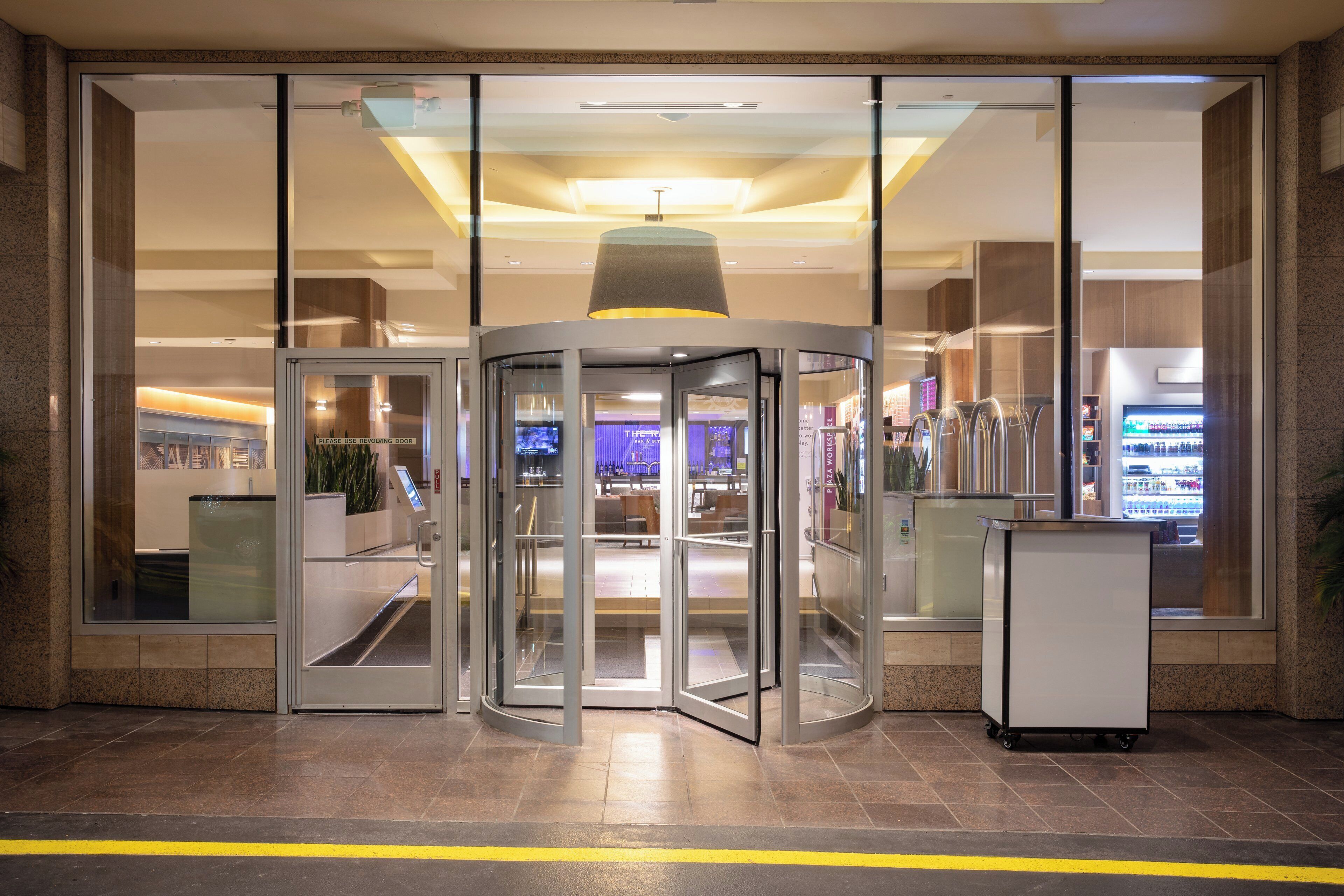 A revolving door entrance to a building with a glass facade, a ceiling light fixture, and a vending machine to the right.
