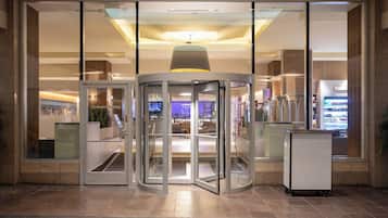 A revolving door entrance to a building with a glass facade, a ceiling light fixture, and a vending machine to the right.