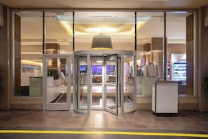Entrada con puerta giratoria a un edificio de fachada de vidrio, una lámpara en el techo y una máquina expendedora a la derecha.