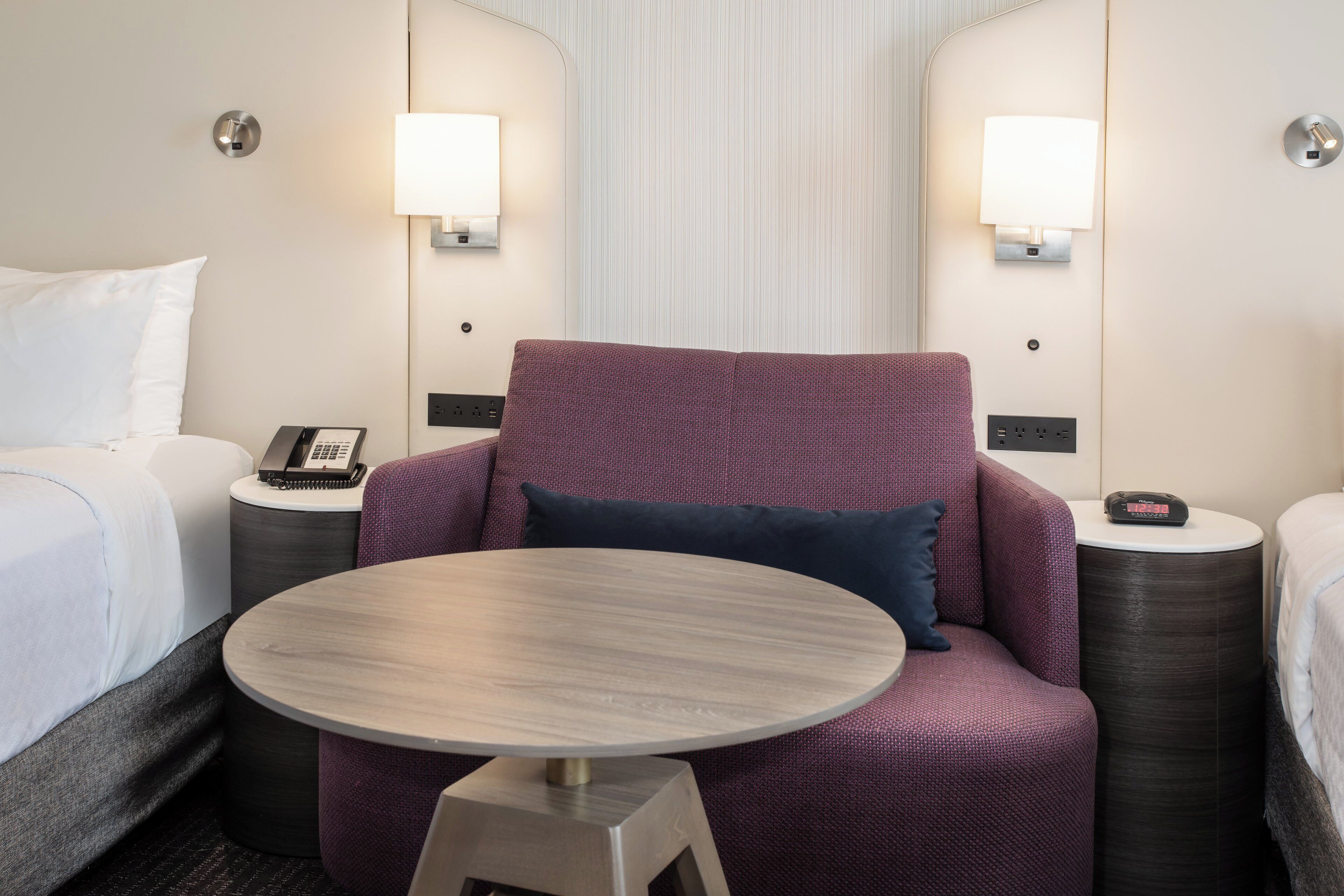 A hotel room with a single bed, a round table, a purple armchair with a pillow, two bedside tables with lamps, and a wall-mounted clock.