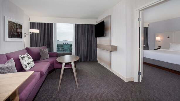 A hotel room with a purple sofa, a small round table, a bed, a TV mounted on the wall, and a view of the city through large windows. - Crowne Plaza Kansas City Downtown by IHG (Kansas City)