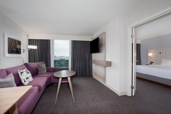 Quarto de hotel com sofá roxo, uma pequena mesa redonda, cama, TV fixada na parede e vista da cidade através de amplas janelas.