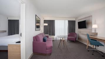 Une chambre d’hôtel avec un lit, un canapé violet, un bureau avec une chaise, une petite table et un téléviseur fixé au mur.