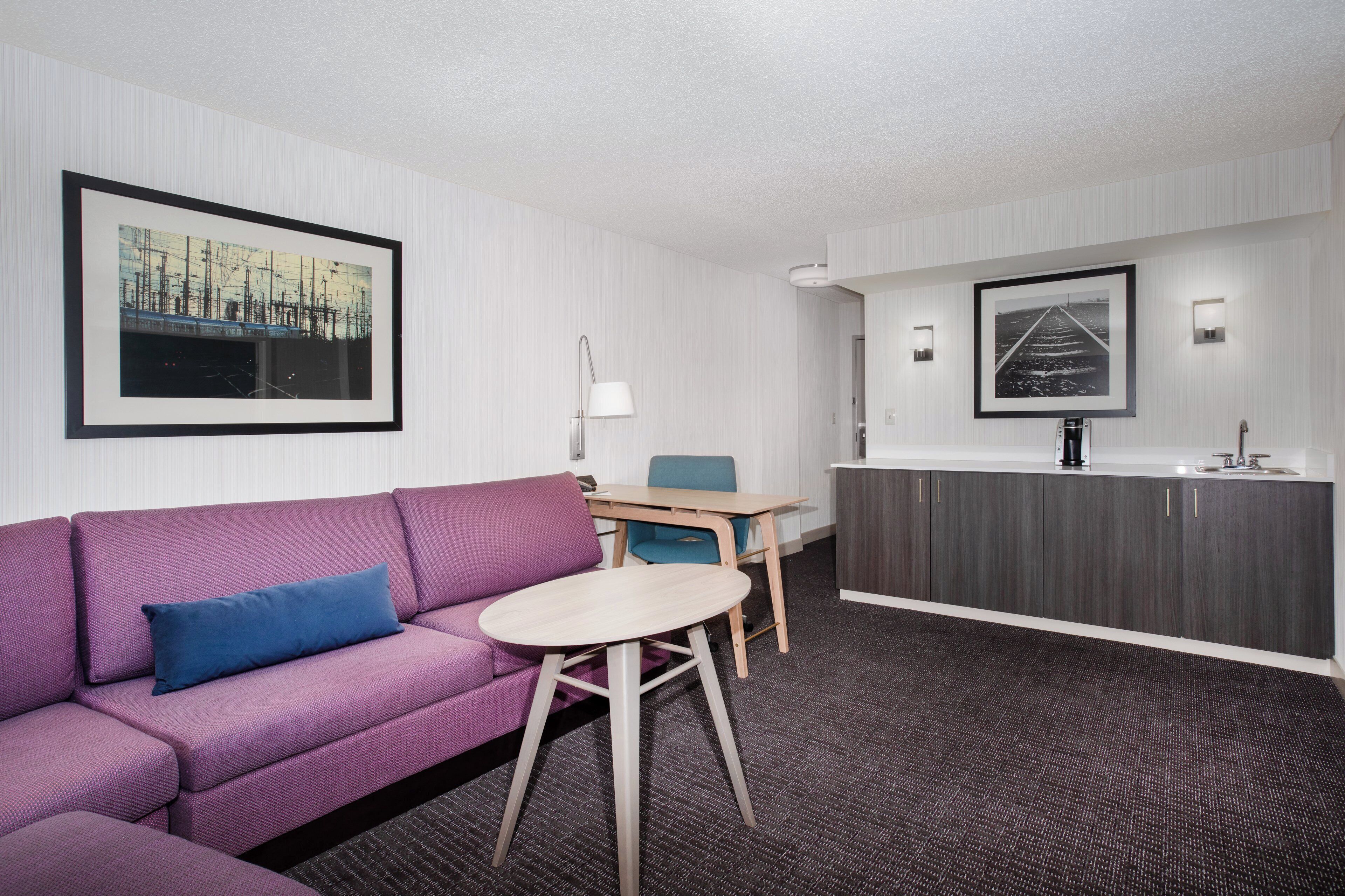 A hotel room with a purple sofa, a small table, a kitchenette, and two framed pictures on the walls.