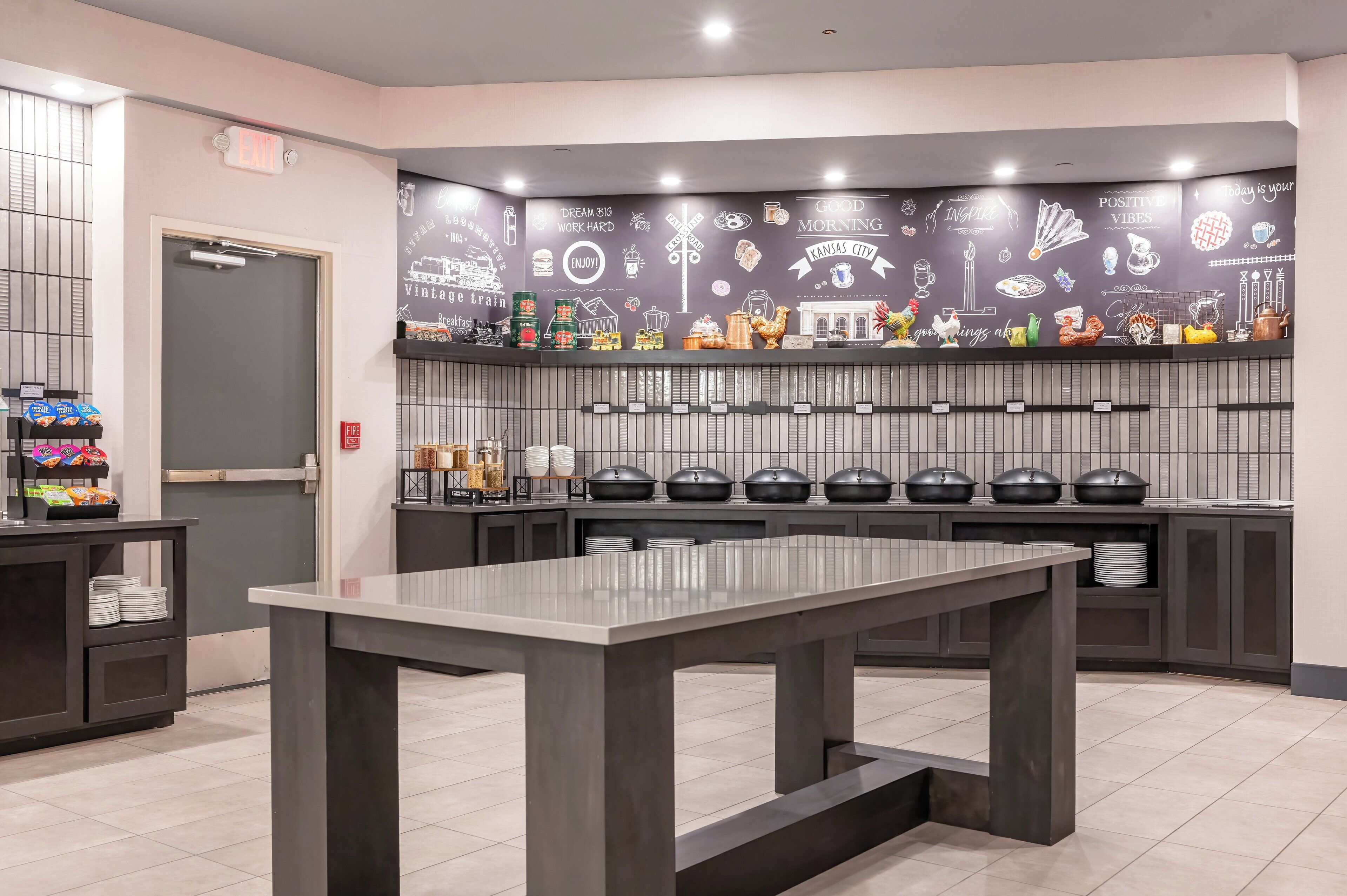 Une cuisine moderne dotée d’un îlot central, d’un mur décoré de diverses illustrations représentant des aliments et des boissons, et d’un buffet avec plusieurs points de service.
