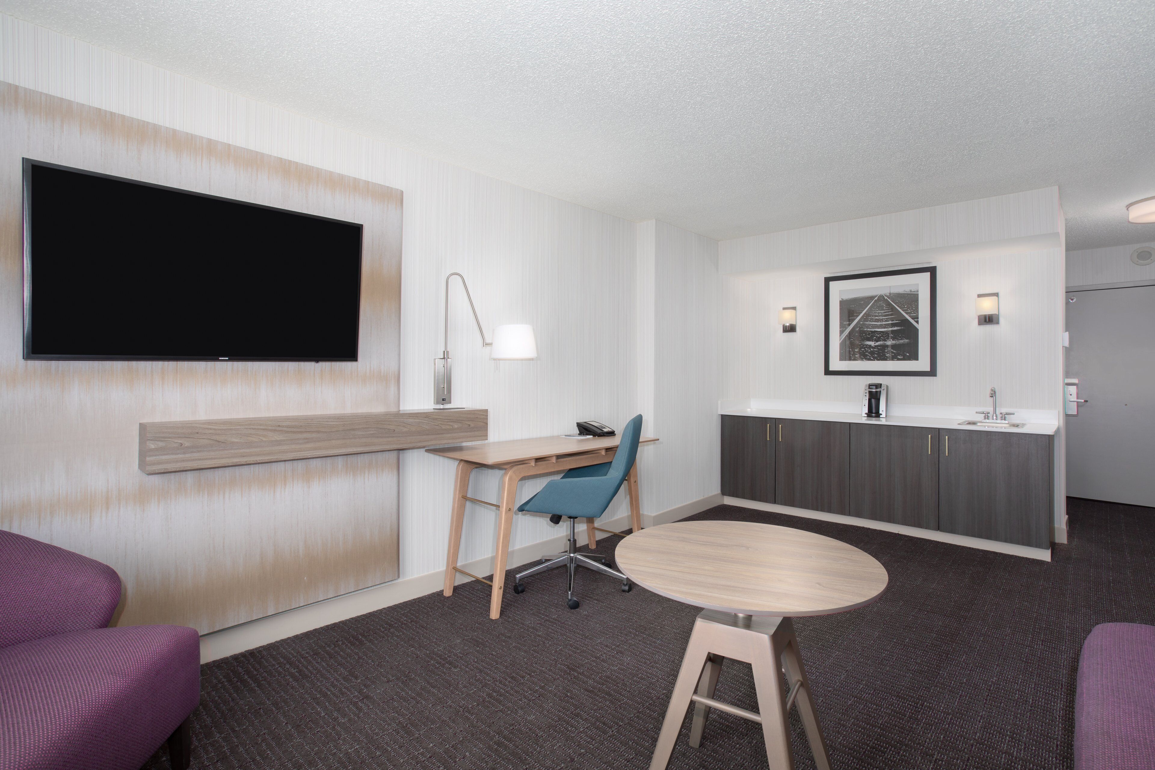 Una habitación de hotel moderna con una televisión de pantalla plana, un escritorio con silla, una mesita redonda pequeña, un sofá púrpura, una kitchenette y un cuadro enmarcado en la pared.