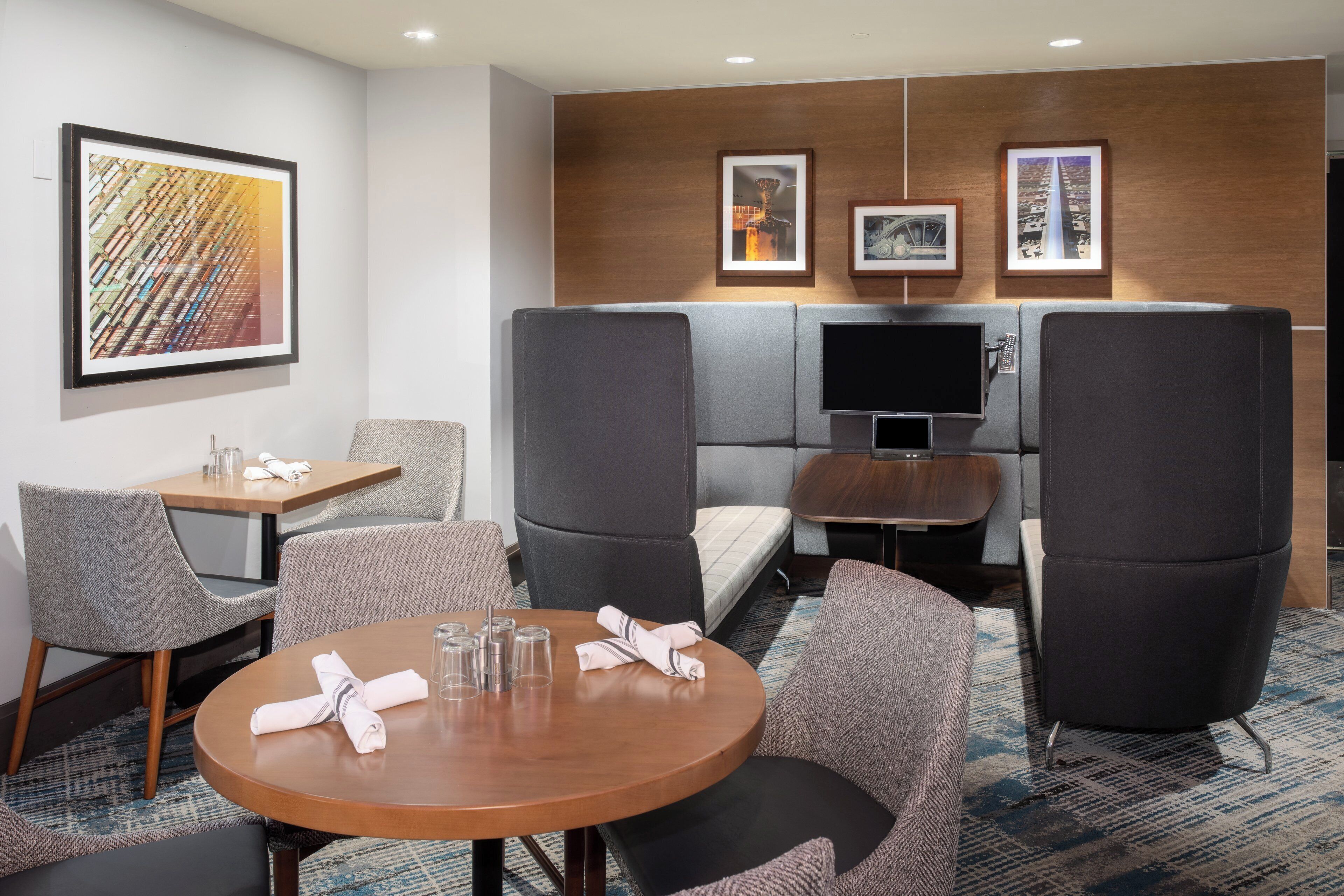 Quarto de hotel com uma mesa redonda, duas poltronas, um sofá, uma televisão e obras de arte na parede.