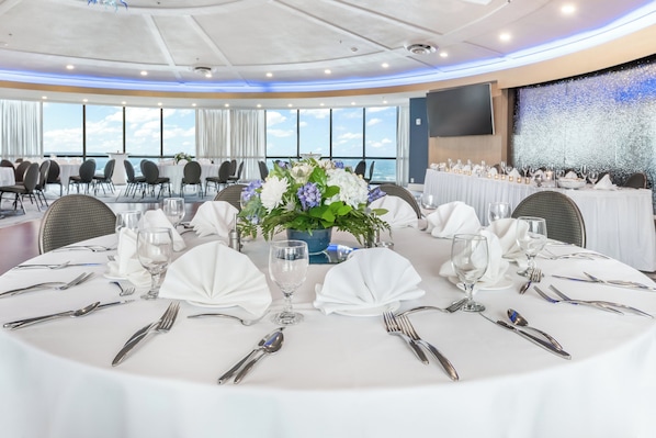 Un salón de conferencias con mesas redondas preparadas para un evento formal, con manteles blancos, cubiertos y centros de mesa florales.