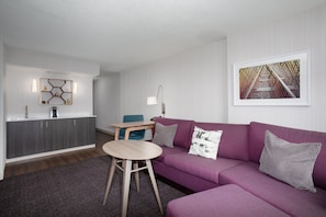 Una sala de estar moderna con un sofá morado, una mesa de centro de madera, una zona de cocina con encimera blanca y un cuadro en la pared.