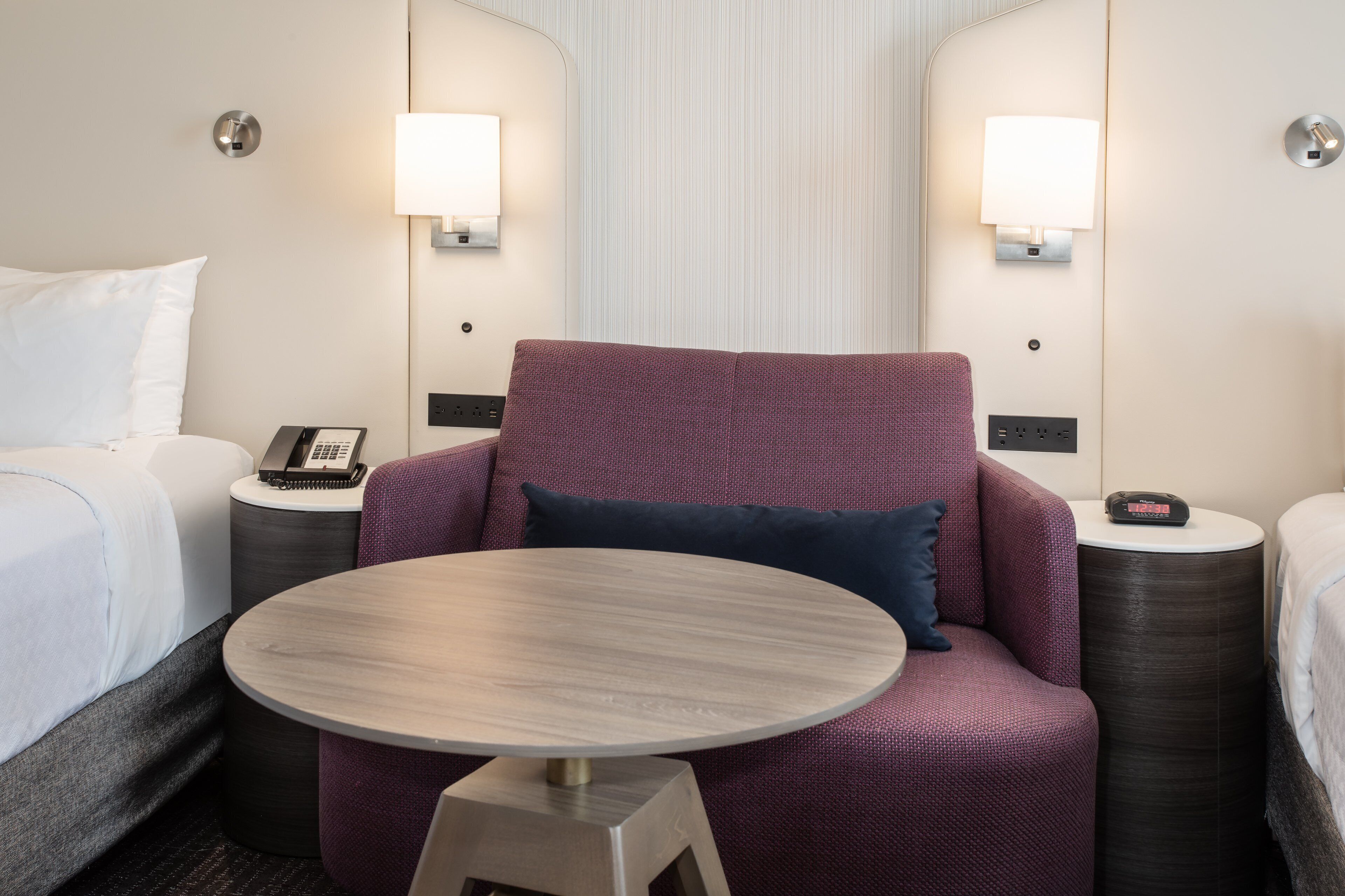 A hotel room with a bed, a round table, a purple armchair, and two bedside tables with lamps.