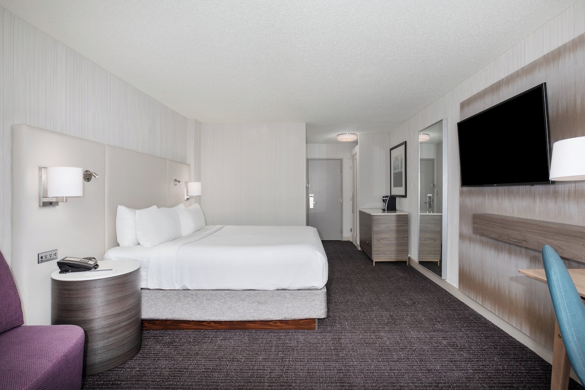 Una habitación de hotel con cama, mesita de noche, lámpara, televisor y una silla.