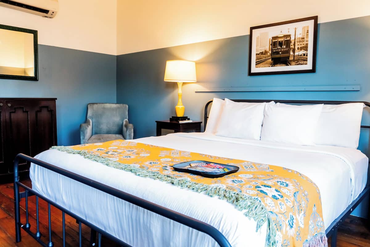 Cama bem arrumada com manta estampada, uma cadeira, mesa de cabeceira com abajur e quadro na parede.