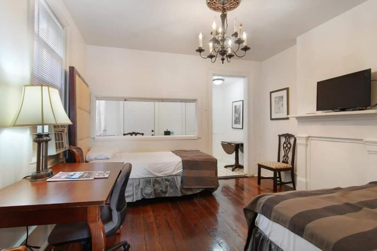 Quarto de hotel com escrivaninha de madeira, cama com colcha listrada, televisão na parede e um candelabro.