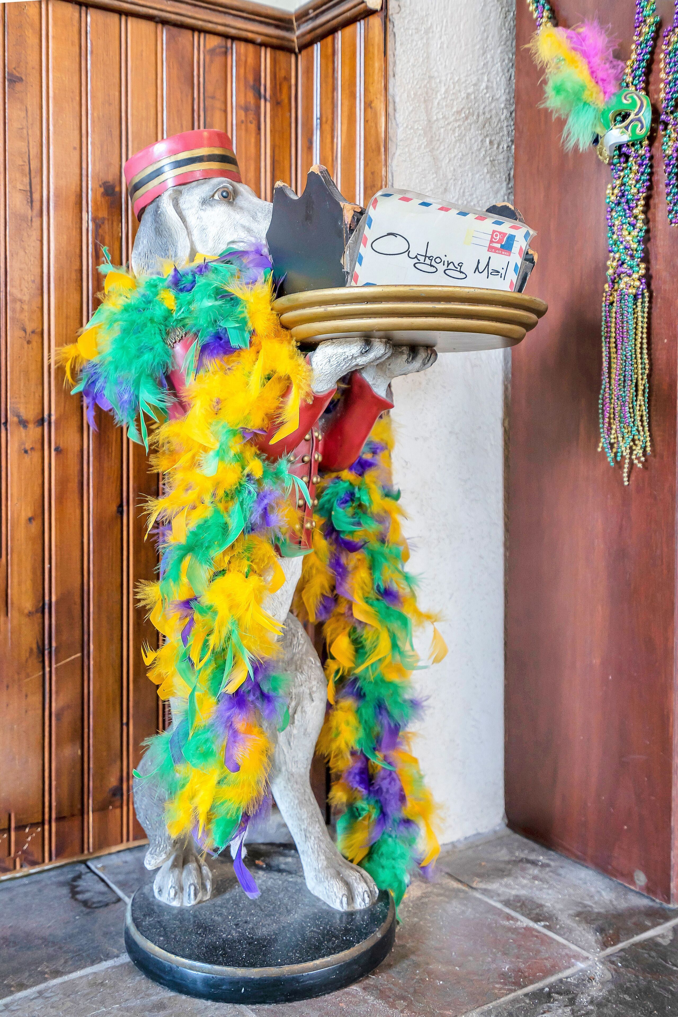 A statue of a dog wearing a colorful feathered headdress and a red sash, holding a tray with a 'Outgoing Mail' envelope.