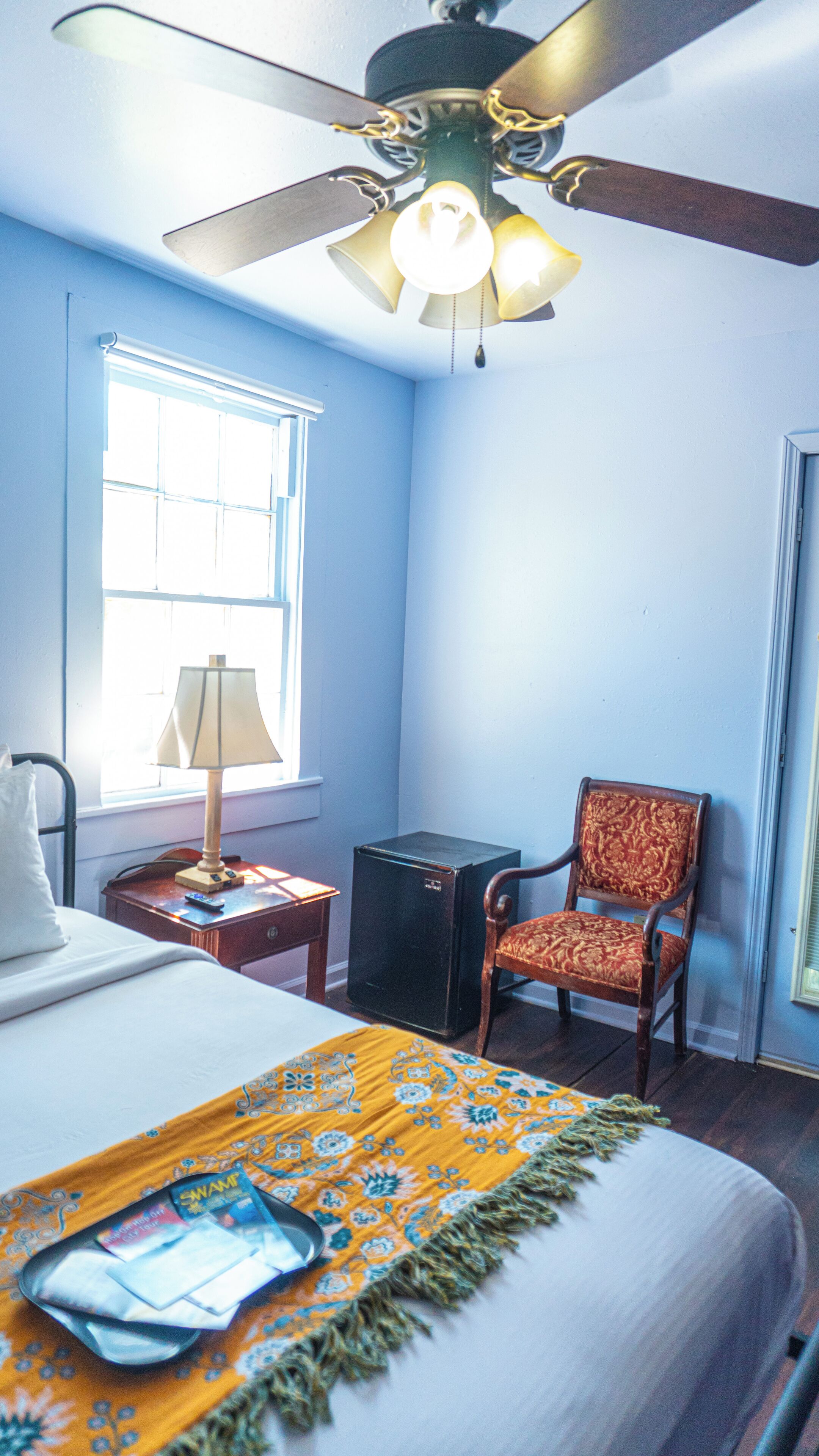 A hotel room with a bed, a chair, a desk with a lamp, a suitcase, and a ceiling fan.