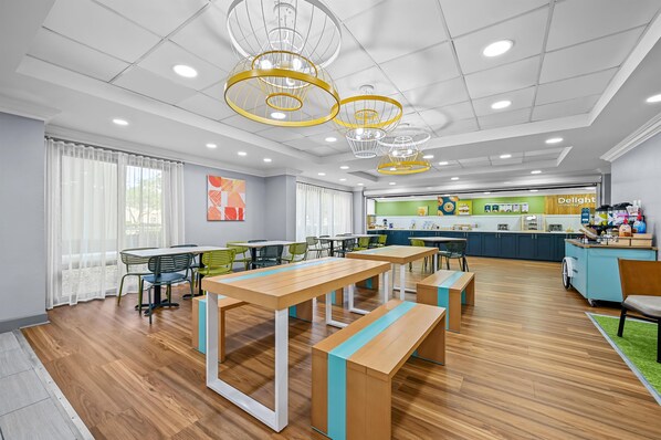 A modern dining area with wooden tables and benches, a kitchen in the background, and a ceiling with recessed lighting and decorative fixtures.