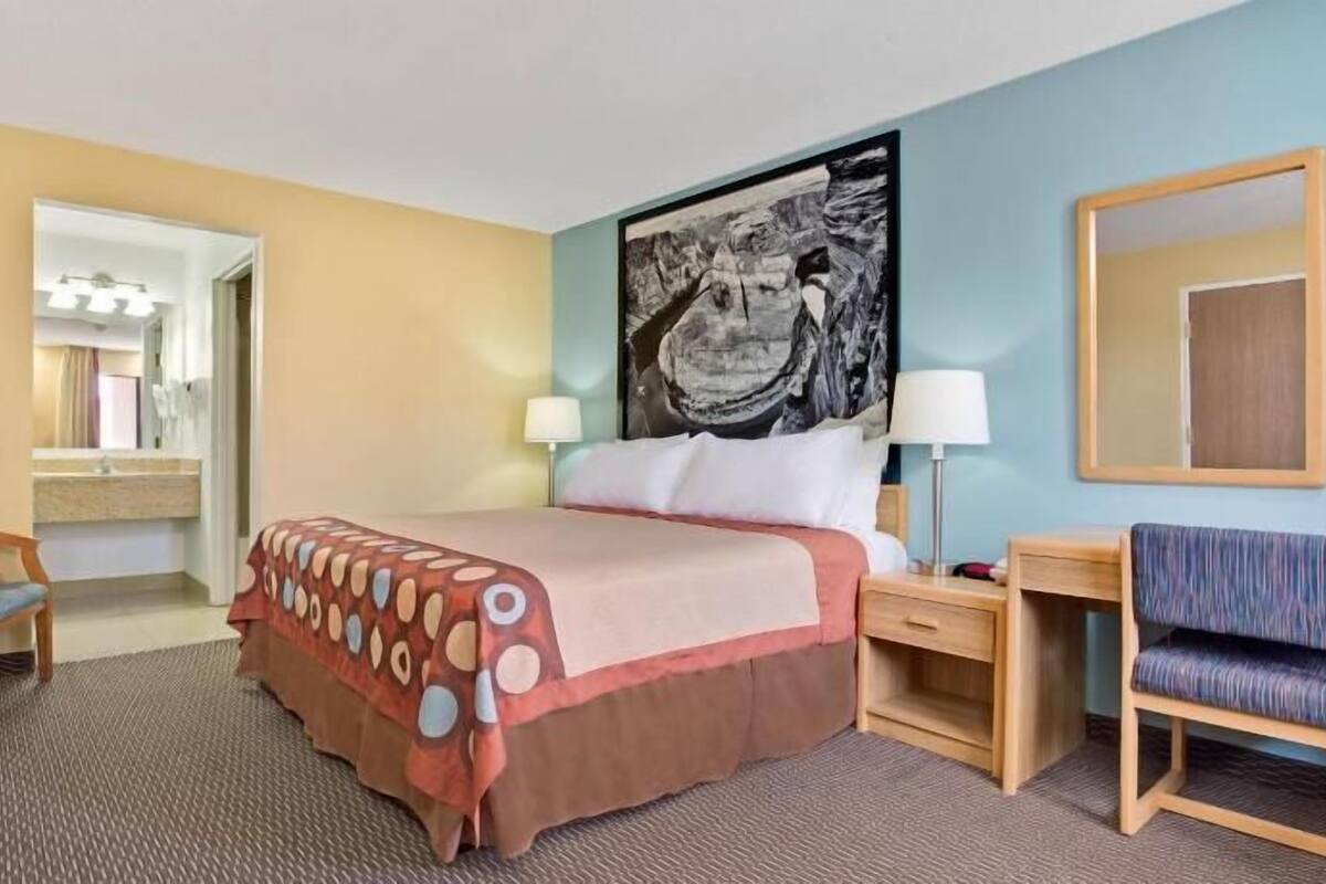 A hotel room with a bed, a desk, a chair, a television, a painting on the wall, and a mirror.
