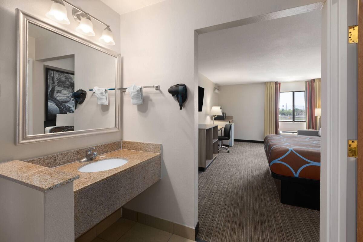 A hotel bathroom with a granite sink, a large mirror, and a hairdryer. The room has a bed, a desk, and a window with curtains.